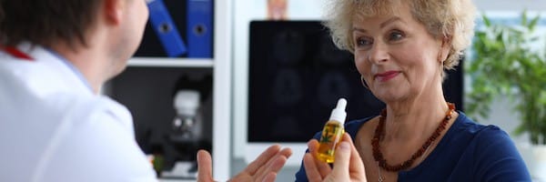 Portrait of aged senior female consulting with doctor. Therapeutic in medical gown advising cannabinoid oil to patient. Practitioner holding bottle with cannabis leaf sign