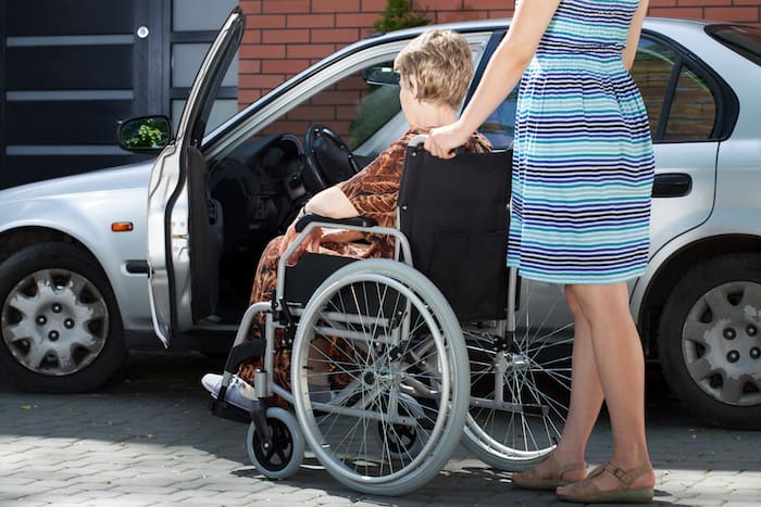 Girl helping senior woman on wheelchair getting into a car, horizontal