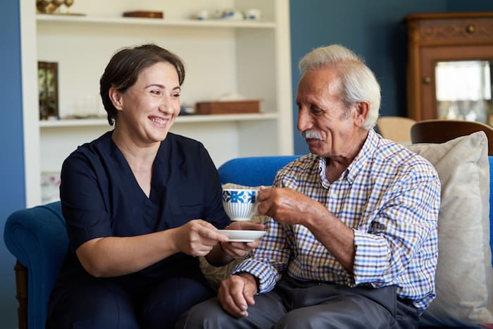 Professional helpful caregiver at nursing home. Health visitor and a senior man during home visit. Young caregiver in uniform and elderly man drinking tea