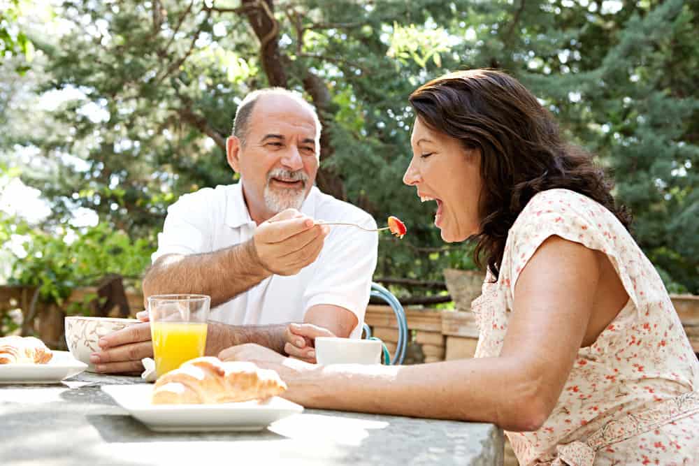Senior couple sitting together having breakfast in a luxury garden during a sunny day on holiday with the husband feeding the wife a strawberry. Mature people eating and drinking healthy food. Outdoors lifestyle.