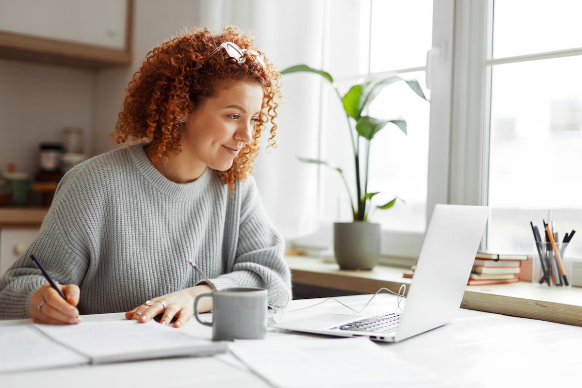 Cute older university student with curly red hair doing homework sitting at kitchen table with coffee cup and wired earphones next to big window, watching online tutorial on laptop and writing in copybook