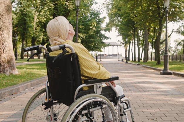 senior woman in a yellow striped shirt sits in a wheelchair outside on a park walkway