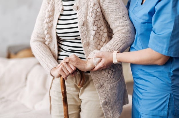 A nurse in blue scrubs helping a senior female patient in a cardigan stand up as she supports herself with a cane.