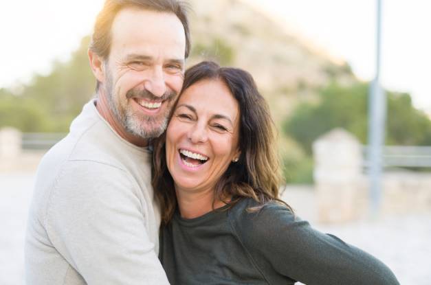 A couple, a man and a woman in their fifties, laughing together and smiling at the camera in a close embrace outdoors.