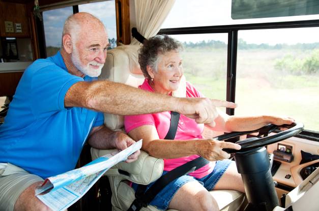 An older retired couple in an RV. The woman is driving while her husband smiles and points to something ahead.