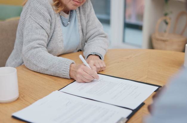 A senior woman signs a contract in an office on a wooden, circular table with two white coffee cups