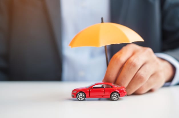A close-up of a man in a suit holding a toy yellow umbrella over a miniature toy red car on a white table.