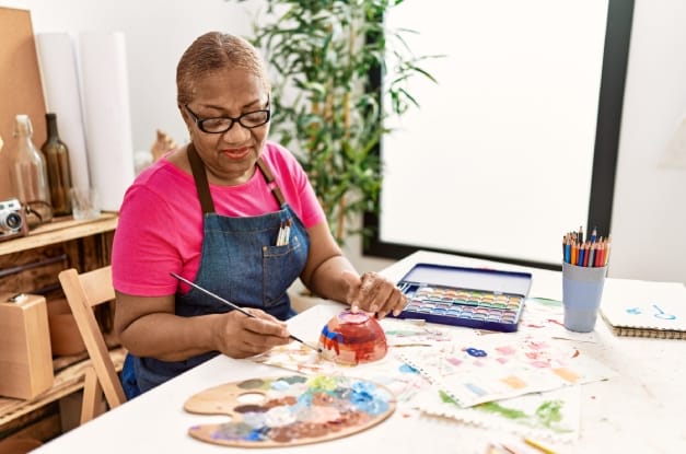A Black senior woman at a craft table enjoying her painting experience. She's wearing a smock and working on a ceramic bowl.