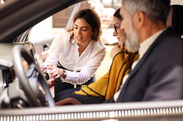 A salesperson in a showroom demonstrates the features of a car interior while a couple sits in the front seats.