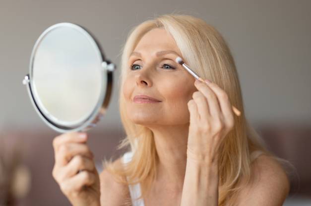 A mature woman with blonde hair smiles as she holds a mirror and uses a makeup brush to apply eyeshadow.