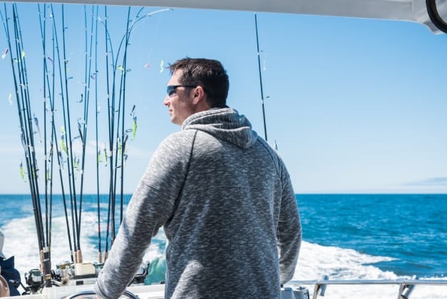 A man wearing a gray hoodie on a boat in the ocean with multiple fishing poles in front of him waiting to be cast.