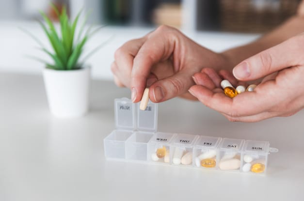 A close-up view shows someone filling a clear pill organizer. There are several pills in each compartment.