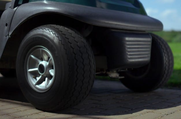 The front two tires of a golf cart, parked on a stretch of pavement, next to lush grass and trees in the distance.