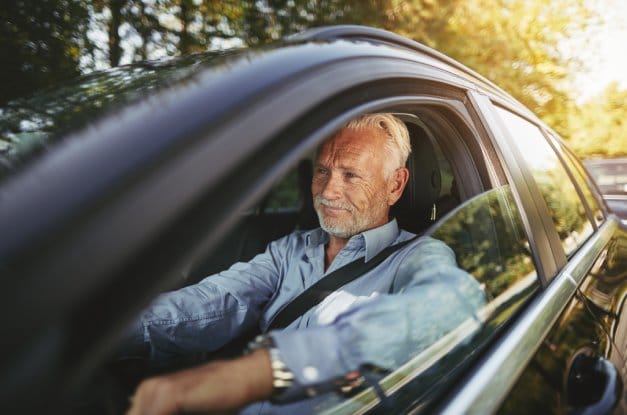 A senior man wearing a blue button-down shirt smiles while driving. The car window is open, allowing you to see his face.