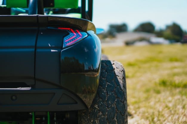 A rear view of a modified, black golf cart with off-road tires and activated LED taillights on a grass field.