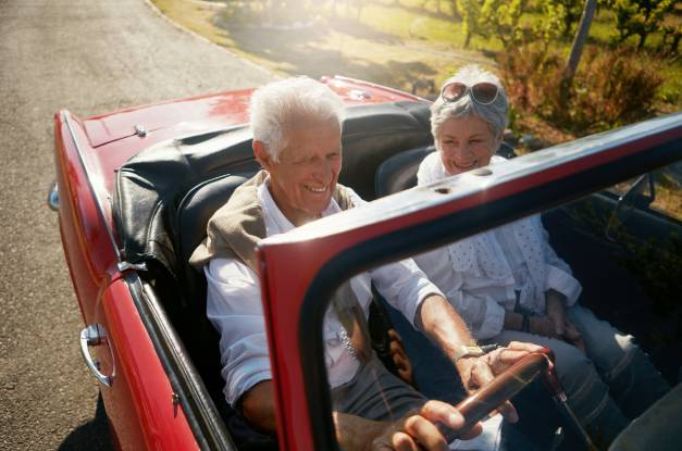 An older couple enjoys a relaxed drive in a red vintage convertible, smiling as they ride together on an open road.