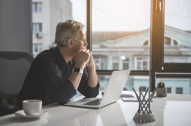 An older man sits at white desk in front of an open laptop and white coffee mug. His head rests on his folded hands.