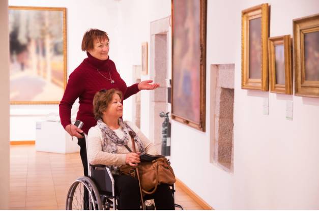 Two older women are looking at paintings in an art gallery. One woman is in a wheelchair and the other stands behind her.