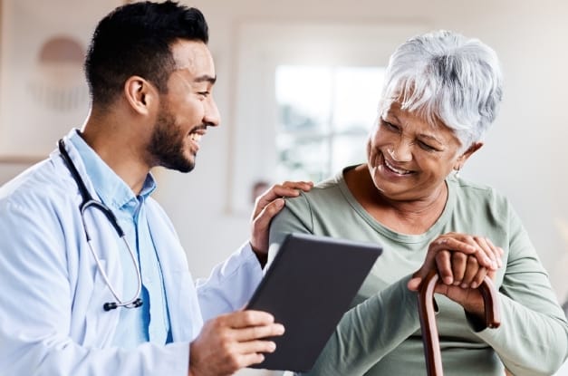 An older adult woman in a green shirt is looking at a tablet with a male doctor who has his hand on her shoulder.