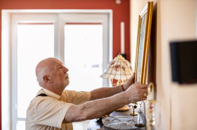A senior man hangs up a framed piece of art on the wall of his home. Sunlight pours in through the glass doors behind him.