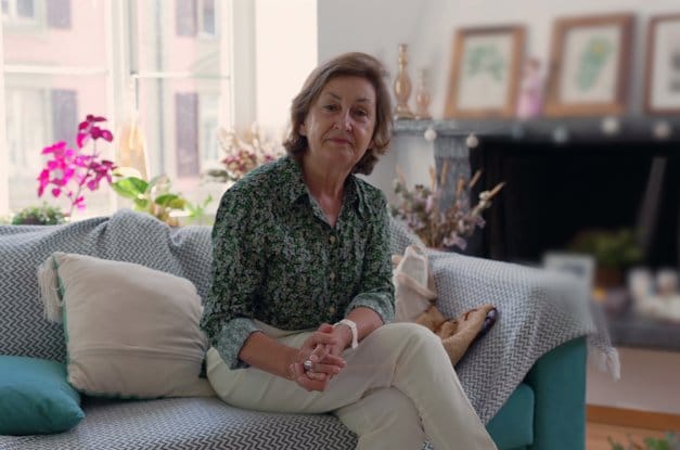 A woman sits on a teal couch covered with a throw and a few cushions. Behind her are flowers, a large window, and a fireplace.