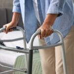 A person gripping the sides of an aluminum mobility walker. They are standing next to a bed with a green blanket.