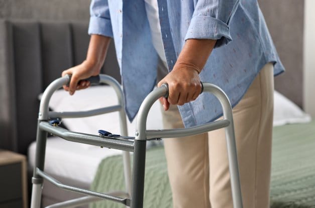 A person gripping the sides of an aluminum mobility walker. They are standing next to a bed with a green blanket.