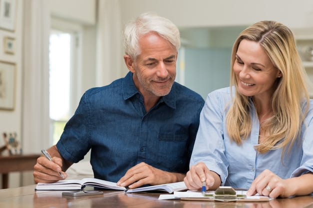 An older couple smiling while reviewing documents. The man is writing in a notebook while the woman holds a pen to a document.