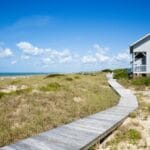 A gray house facing the ocean with a wooden walkway and grassy sand in front of it on a sunny day.