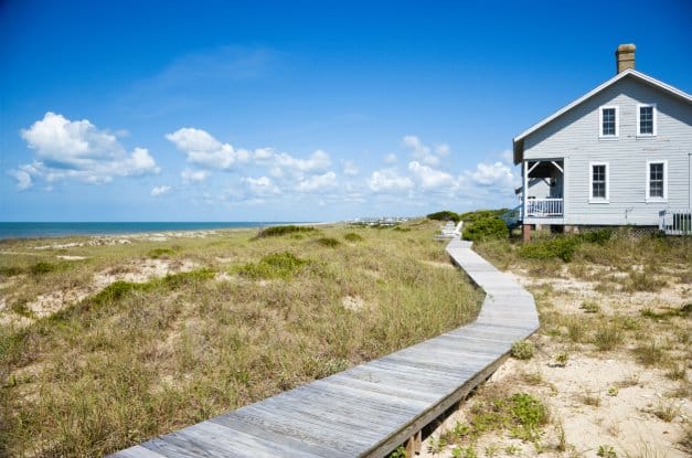 A gray house facing the ocean with a wooden walkway and grassy sand in front of it on a sunny day.