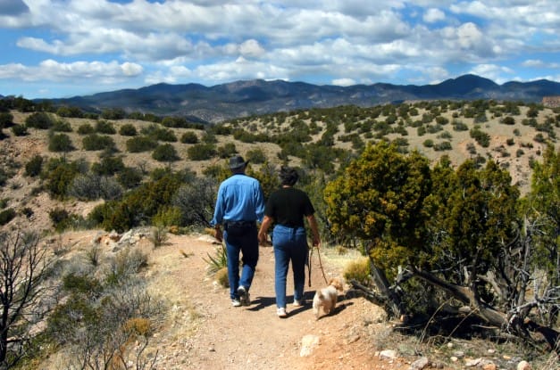 An older man and woman in denim are seen from behind as they hike through a beautiful scrubby landscape.