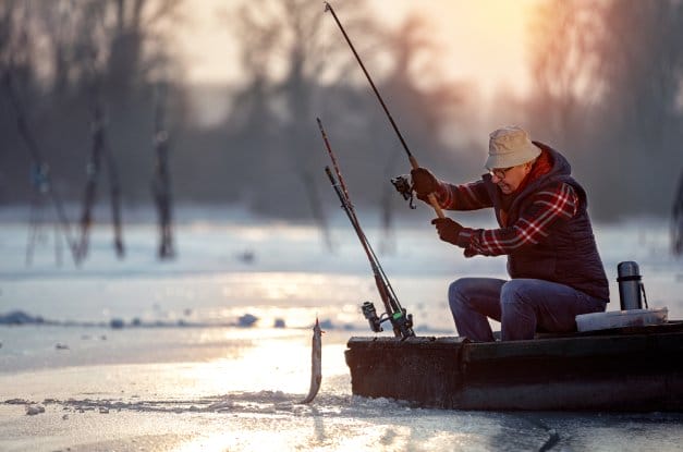 A fisherman wearing a tan hat sitting on his black boat while reeling in a fish out of the water with his fishing pole.