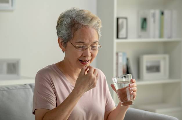 An older woman wearing glasses puts a pill in her mouth. She is holding a glass of water in her other hand.