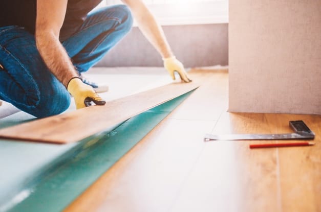 A man wearing yellow work gloves laying a board of laminate faux wood flooring with natural light coming in from the window.
