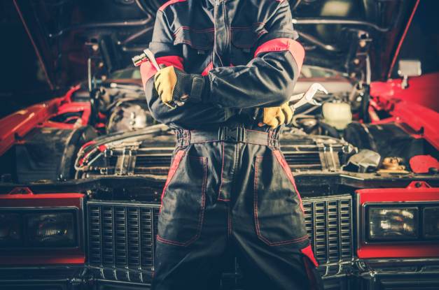 A mechanic in a black and red work suit crosses their arms and leans against a retro car with the engine hood open.