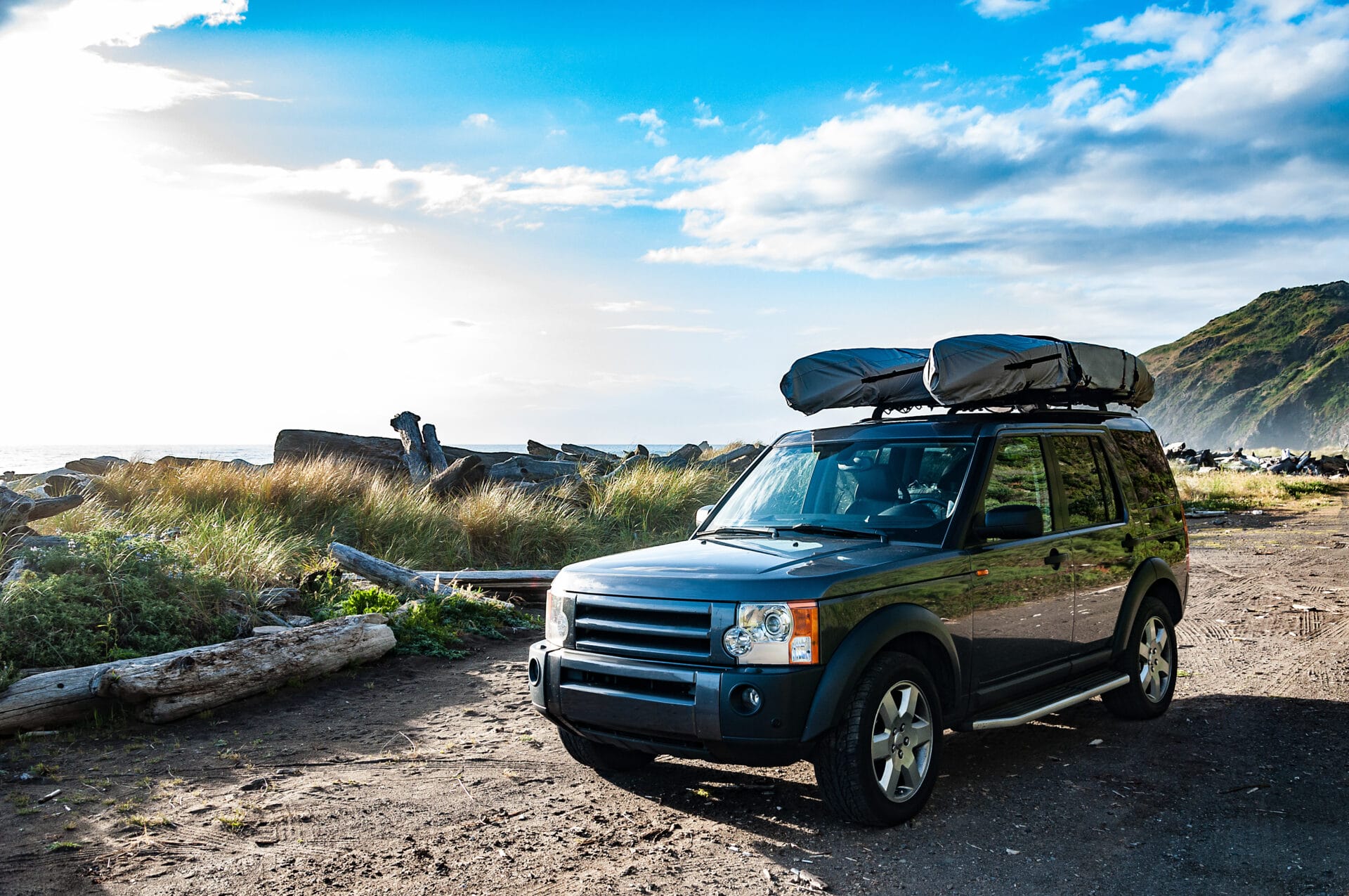 A black SUV with covered kayaks on top is parked on a beach with grassy mountains in the background.