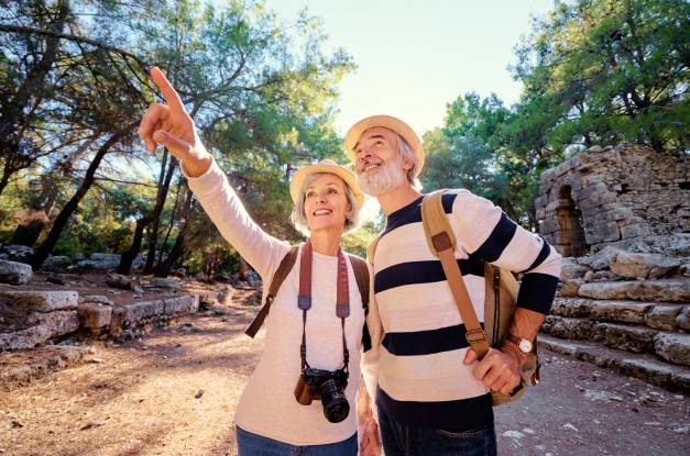 An older woman with a camera around her neck points to something. She and an older man are wearing travel bags and hats.