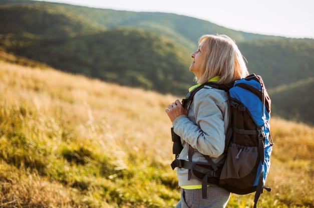 An older adult standing on a grassy hill. She wears a large hiking backpack and a long-sleeved jacket.