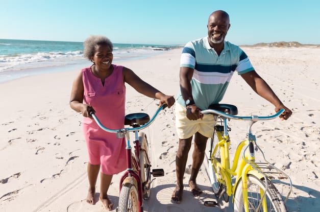 A grinning older Black couple walking bikes on the shore of a white-sand beach on a clear, sunny day.