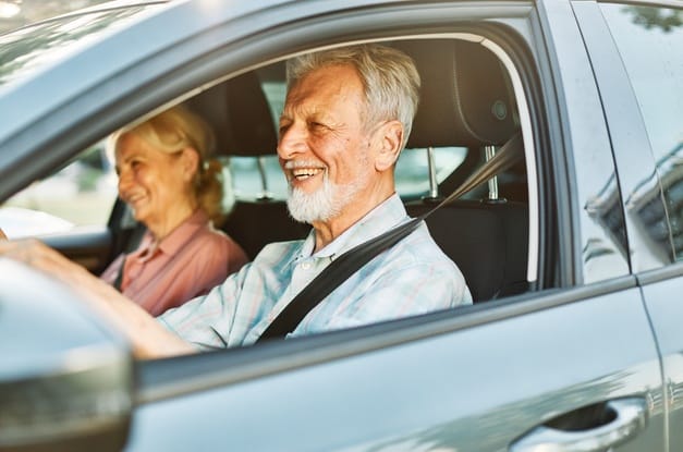 An older man in the front seat of a light gray vehicle smiling as he drives. A woman sits in the seat beside him.