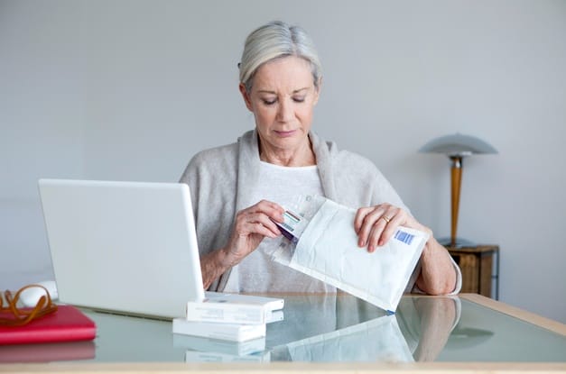 A senior woman sitting at a desk with a laptop on it. She's pulling a box of medication out of an insulated white envelope.
