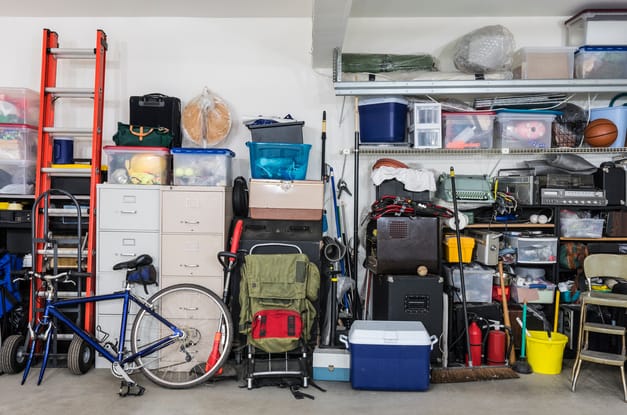 The cluttered wall of a garage. There are full shelves, stacks of storage bins, and various other items against the wall.