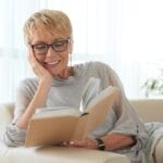 A senior woman wearing readers relaxes on a couch with her head in her hand as she reads a book.
