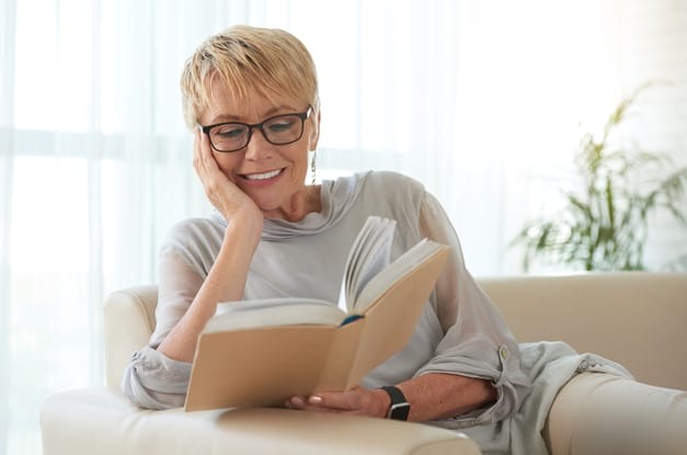A senior woman wearing readers relaxes on a couch with her head in her hand as she reads a book.
