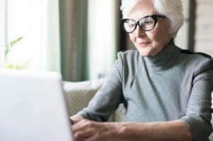 An old woman with white hair wears a gray turtleneck and cat-eye readers as she types on a laptop in a living room.