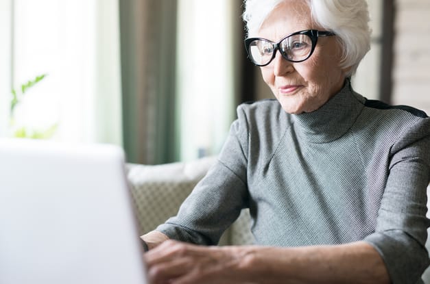 An old woman with white hair wears a gray turtleneck and cat-eye readers as she types on a laptop in a living room.