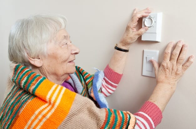 A senior woman with a colorfully striped blanket draped over her shoulders adjusts a thermostat on a beige wall.