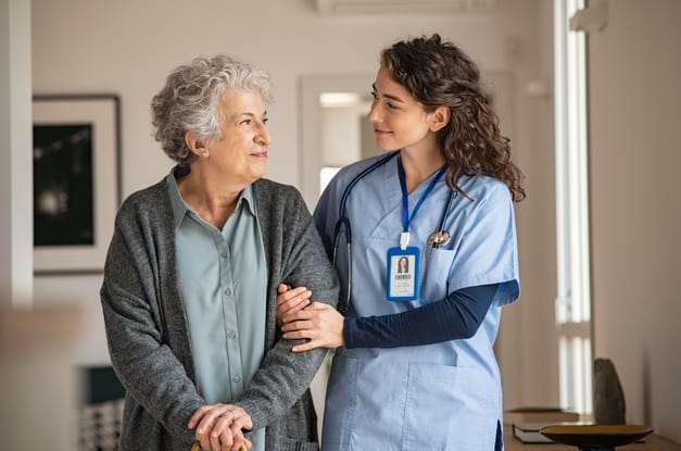 A senior woman with a cane walking next to a younger woman wearing blue scrubs and a stethoscope around her neck.