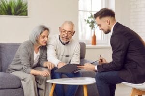 An older adult couple sits on a gray couch, looking at a portfolio that a young businessman is holding.