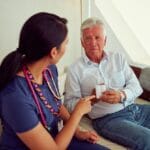 A nurse sitting on the couch with an older gentlemen. She is pointing to a white pack of pills in his hand.